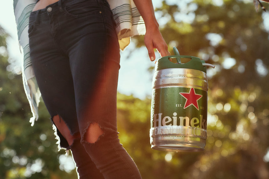 Person holding a Heineken keg outdoors with trees in the background