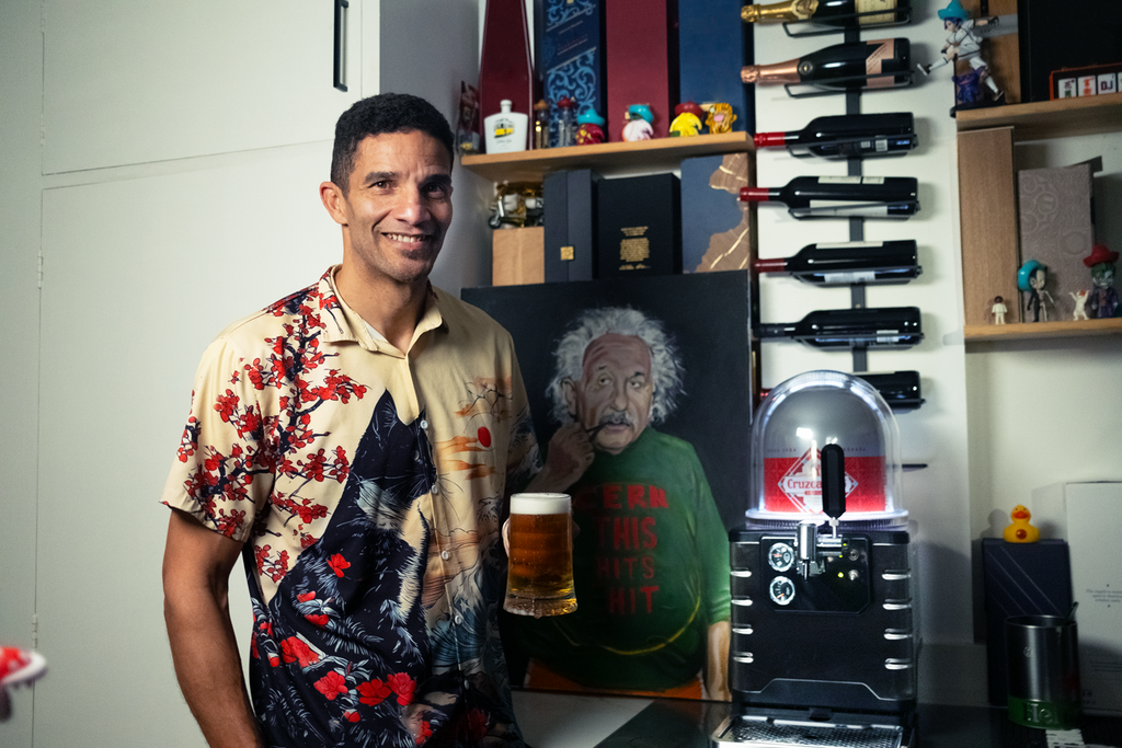 David James holding a beer beside a Blade machine, with a painting of Einstein, shelves, and wine bottles in the background.