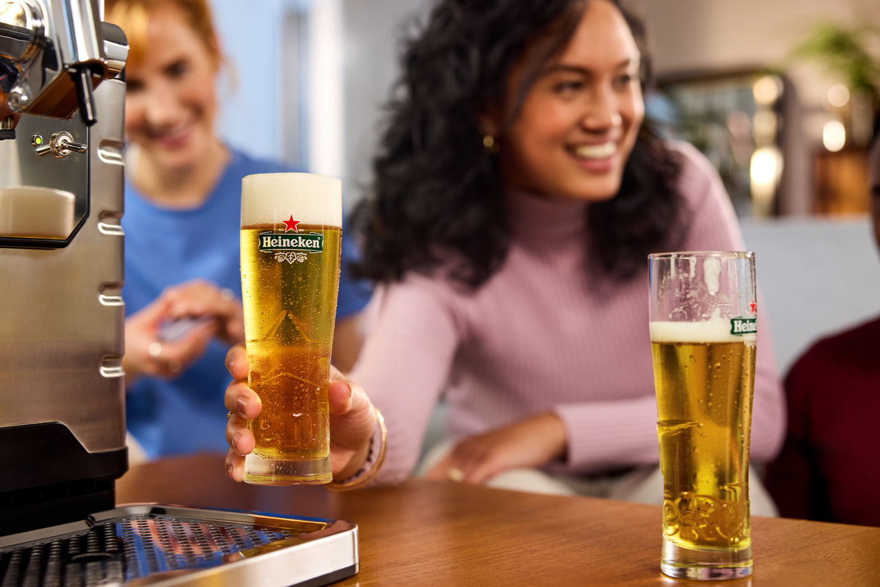 A woman holding a glass of beer with Heineken logo, and another Heineken beer glass on a bar counter