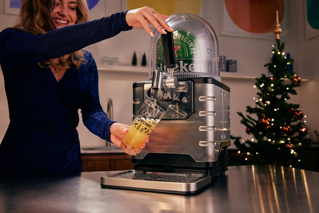Woman pouring a glass of beer from Heineken beer dispenser in a kitchen setting with a Christmas tree in the background.