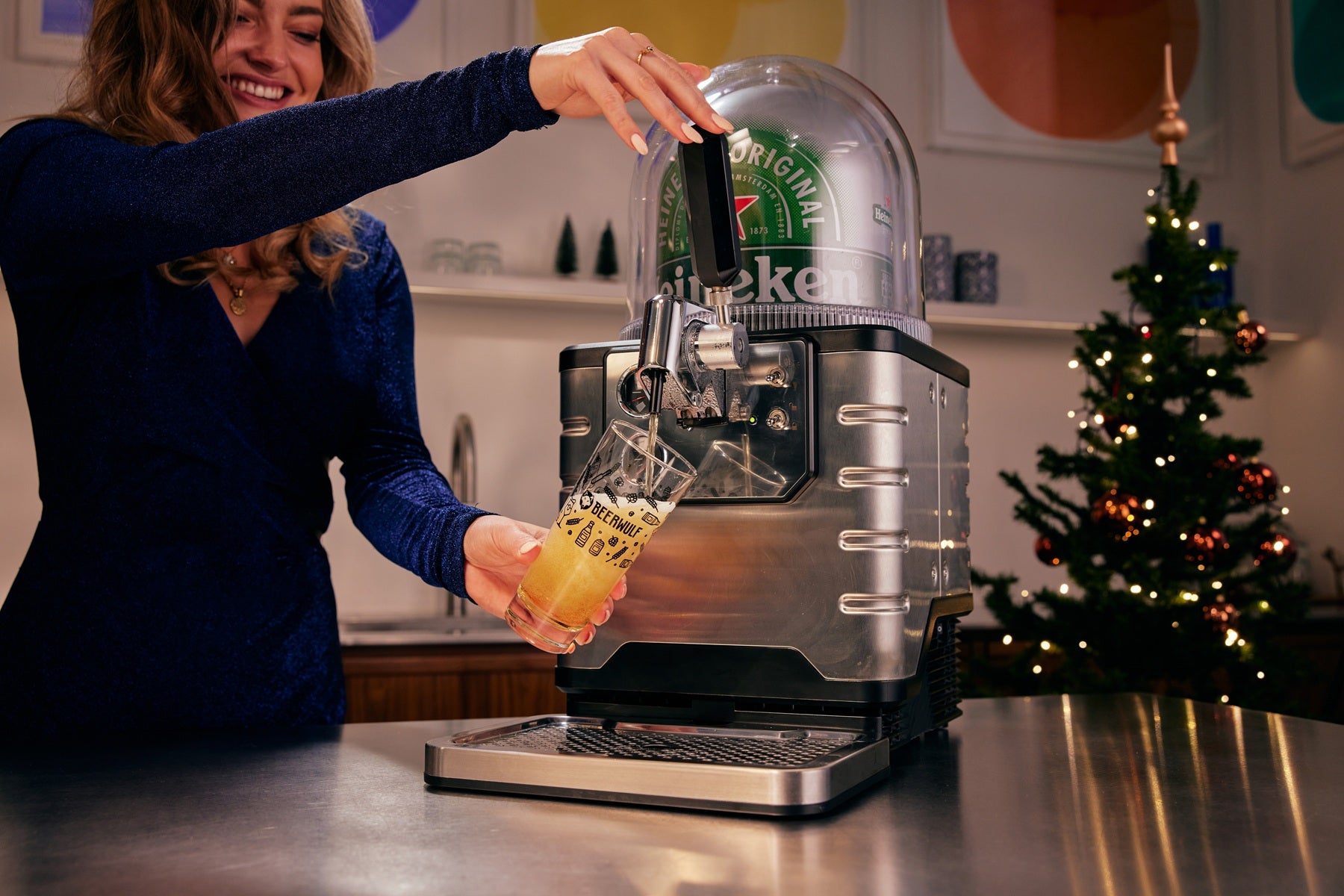 Woman pouring a glass of beer from Heineken beer dispenser in a kitchen setting with a Christmas tree in the background.