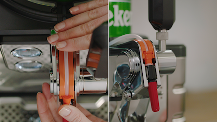 A person inserts a beer keg vertically into a cooling bowl of a machine, aligning the tube at the front. The keg’s tube is fitted into an orange groove, and the black valve is pressed into a metallic holder. The tap cover is flipped up and locked in place, and the red cap is removed.