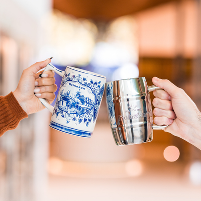 Two hands holding beer mugs in a toast. One mug is white ceramic with blue details. The other is steel.
