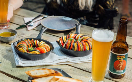 An outdoor table with two pans of grilled vegetables, bread, olive oil and a glass of beer.